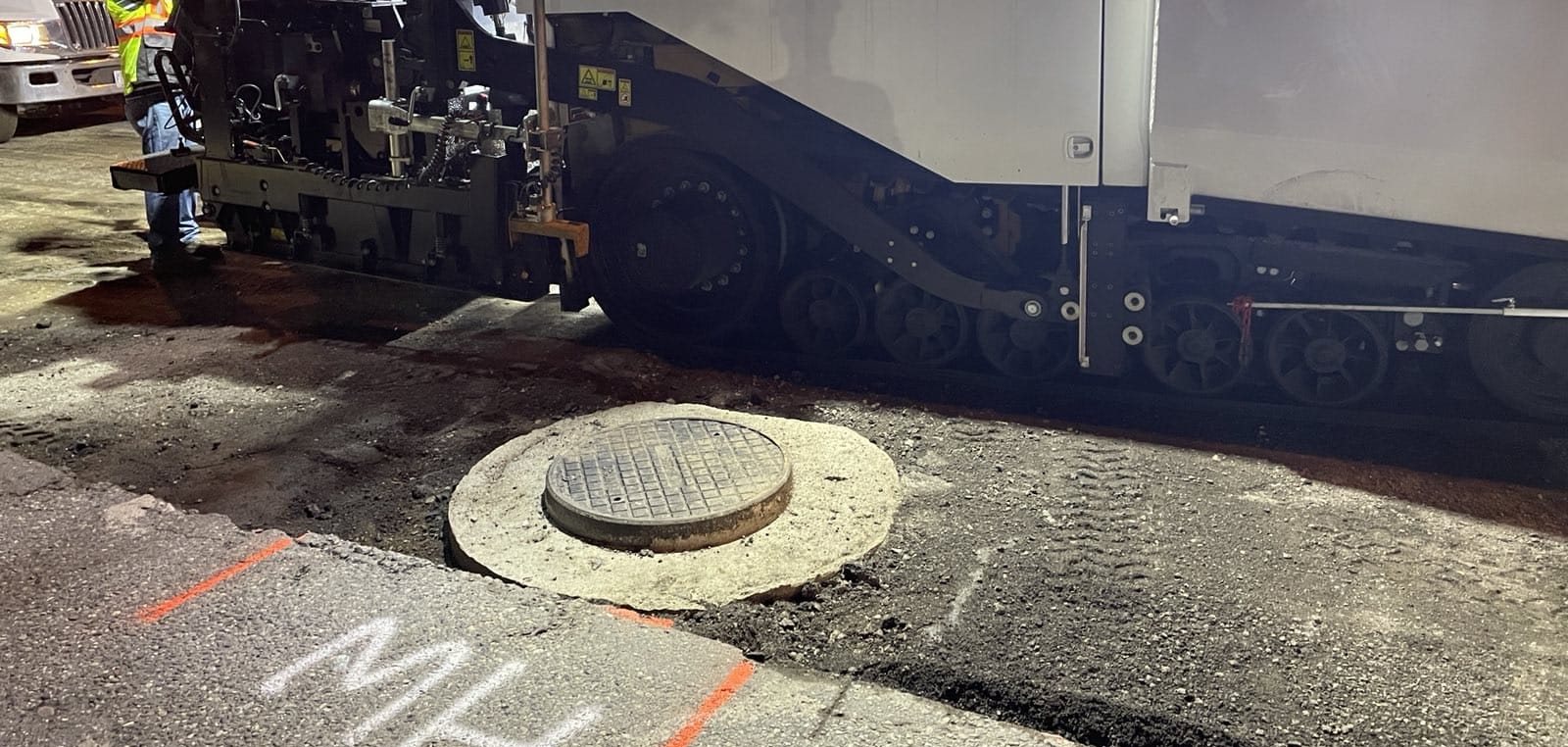 Finished manhole collar in the road with a paving machine working beside it at night